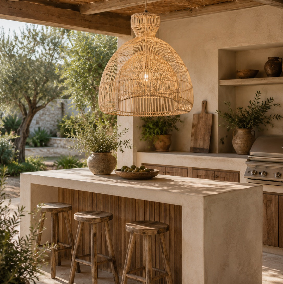 A large woven rattan pendant light hangs over a rustic outdoor kitchen island with three wooden bar stools, set under a covered patio with olive trees in the background.