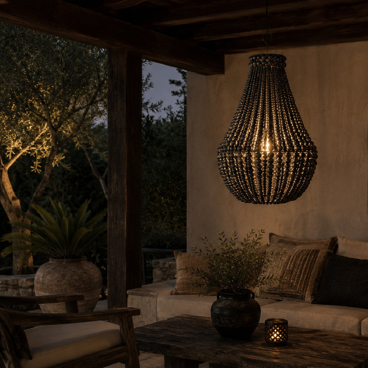 A dark woven beaded chandelier hangs over a rustic outdoor patio seating area at dusk, with a sofa, table, plants, and warm ambient lighting in the background.