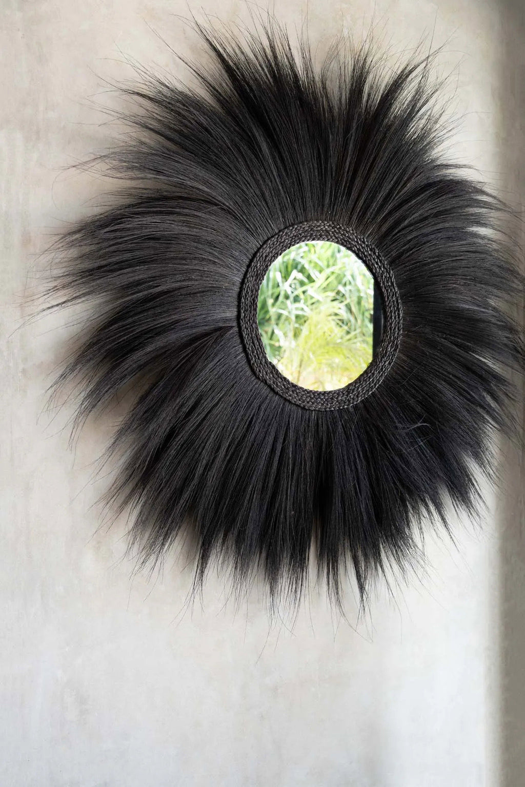 A round black grass wall mirror with a beaded circular frame hangs on a warm beige plaster wall above a stone sink in a rustic, sunlit kitchen.